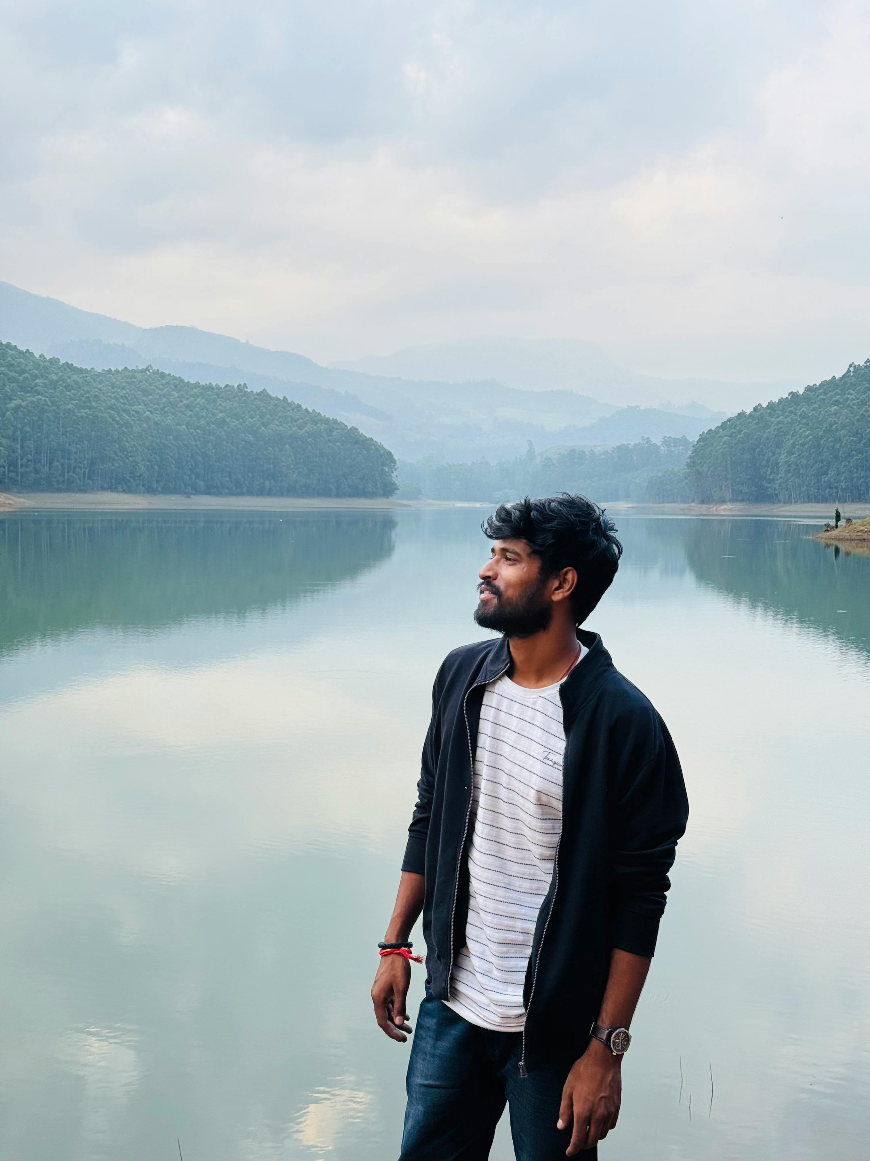 A young man stands by a calm lake with mountains and forests in the background, reflecting tranquility.