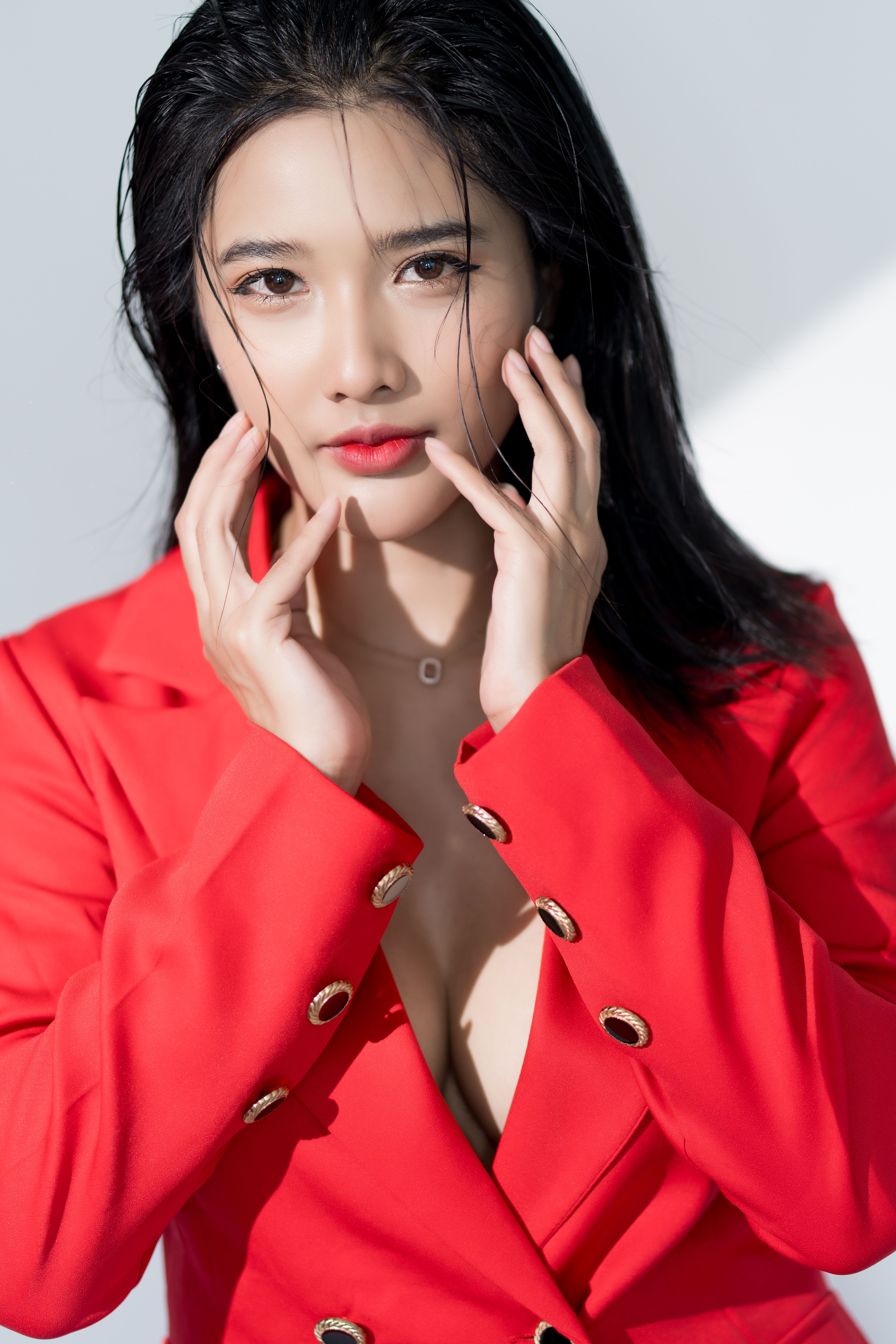 Fashion portrait of an Asian woman in a red suit jacket posing with hands on cheeks in a studio setting.