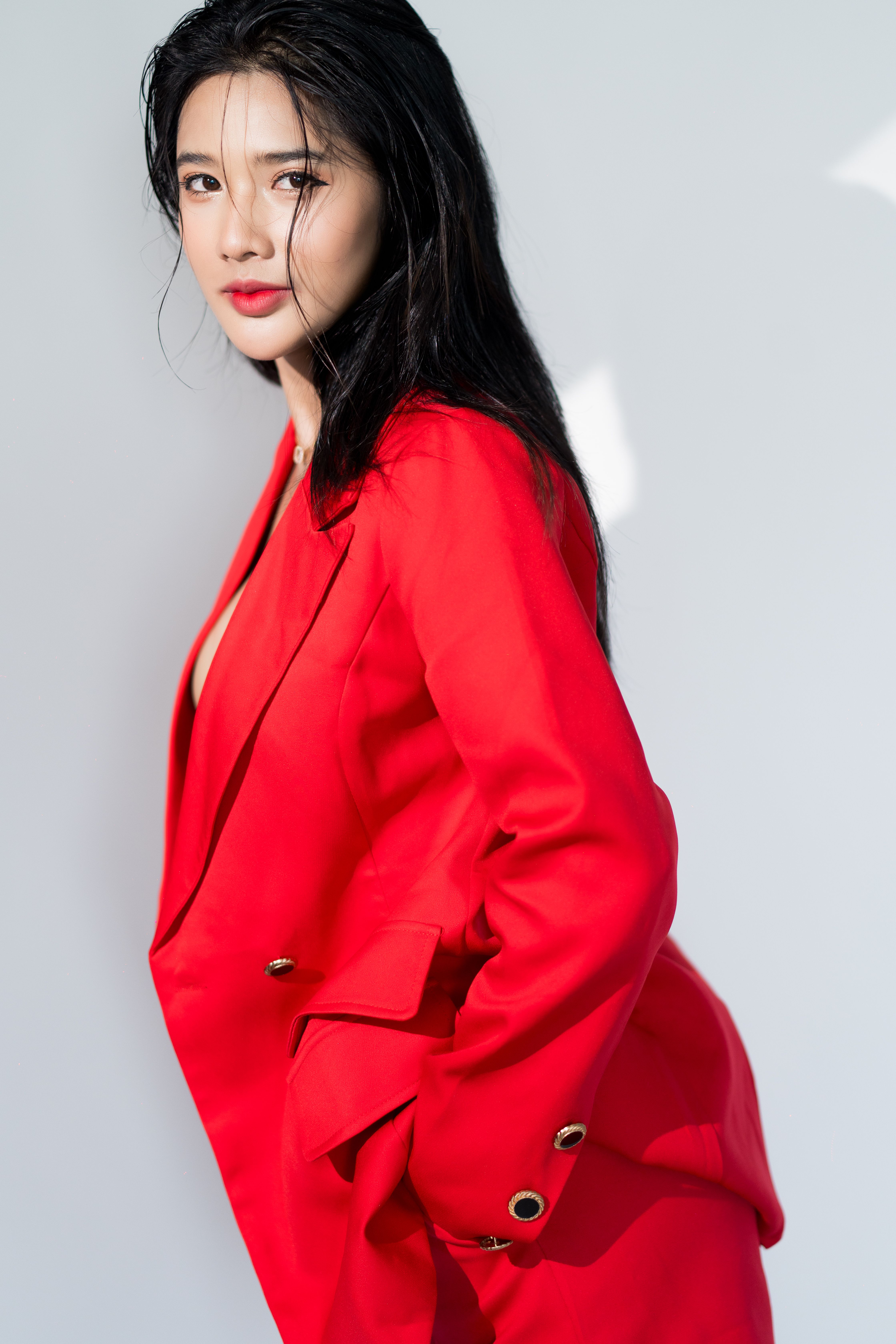 A fashionable portrait of a woman with long black hair in a red suit jacket, set against a white background.