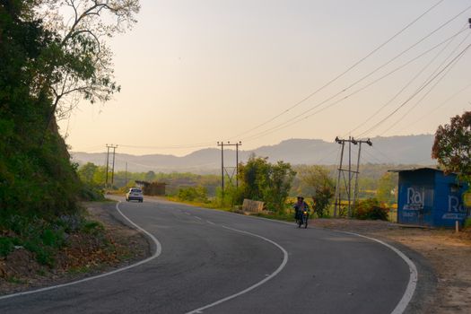 Serene countryside road with a motorbike and car during a peaceful sunset, surrounded by hills.