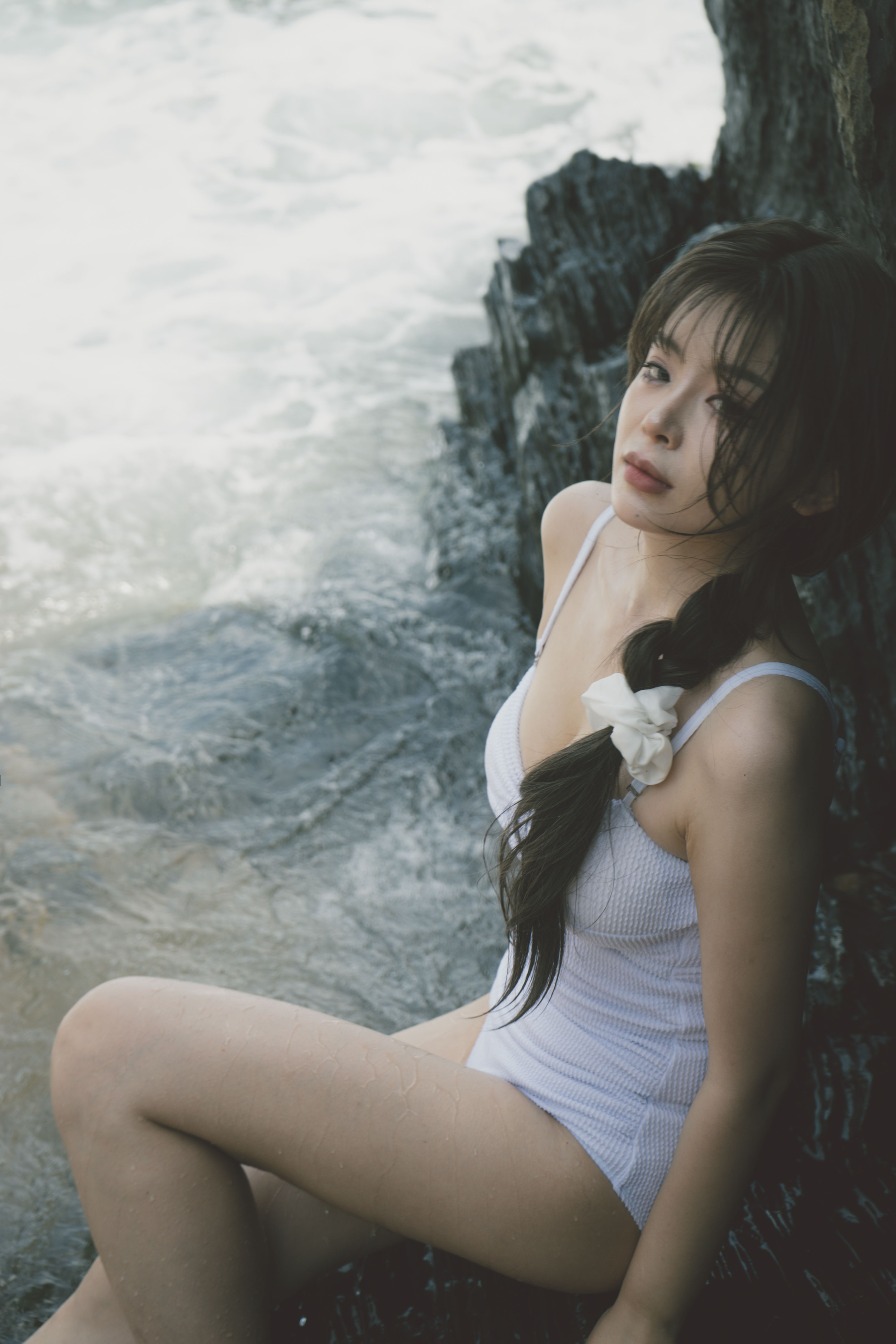 Asian woman relaxing by the ocean in a white swimsuit, sitting on rocks.