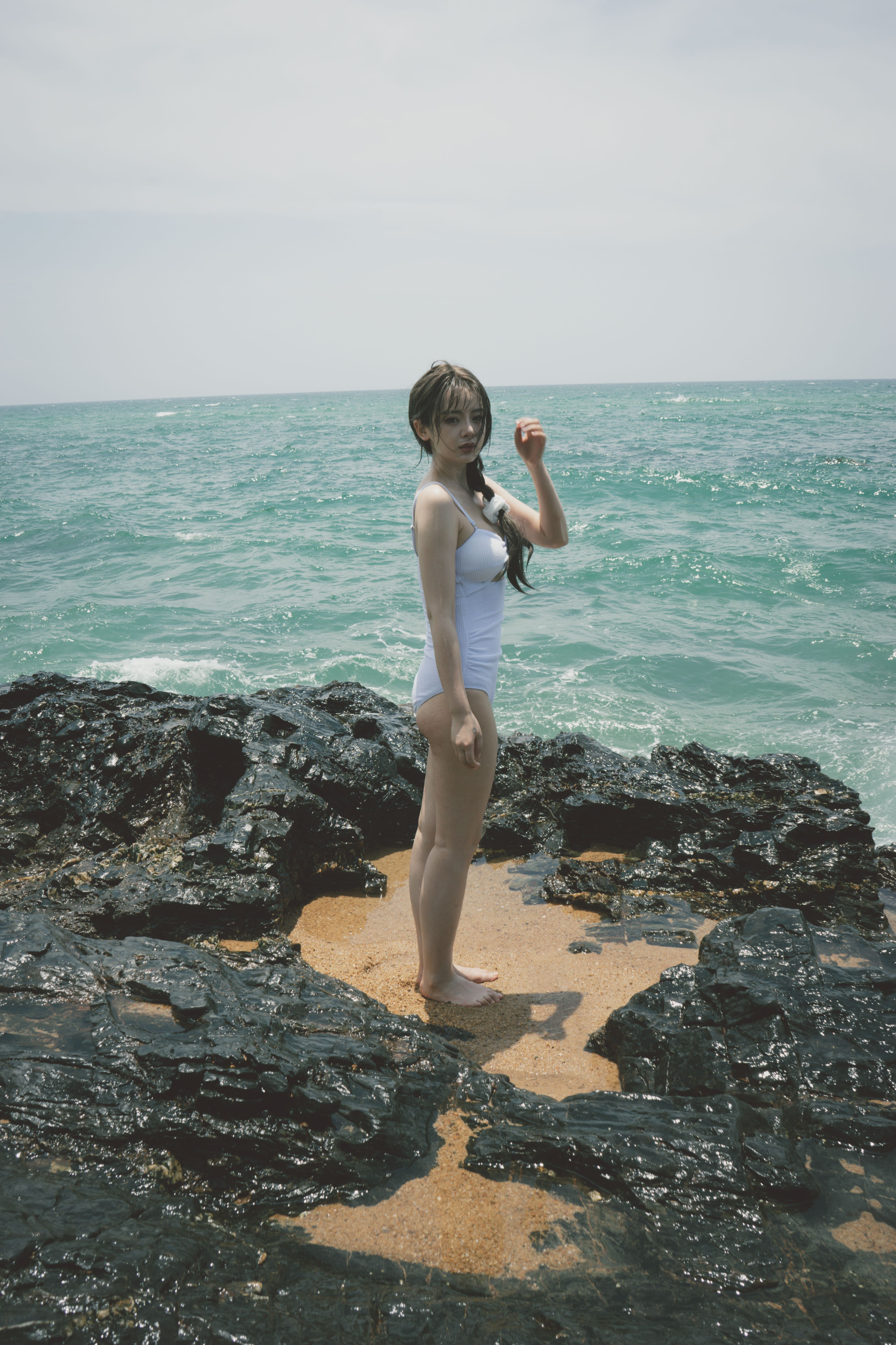 Asian woman in white swimsuit standing on rocky shore by the ocean.