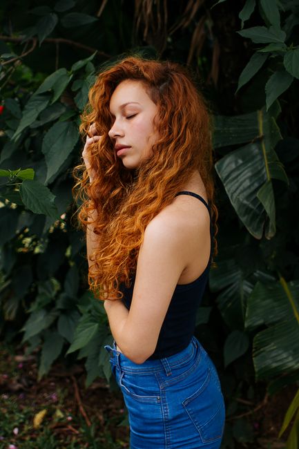 Side profile portrait of a beautiful woman with red curly hair standing against vibrant foliage.