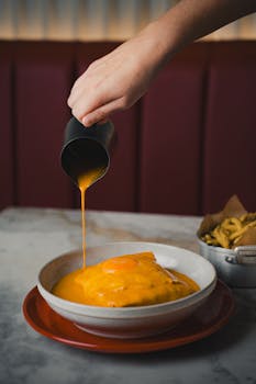 Hand pouring cheese sauce over a dish in a casual dining setting.