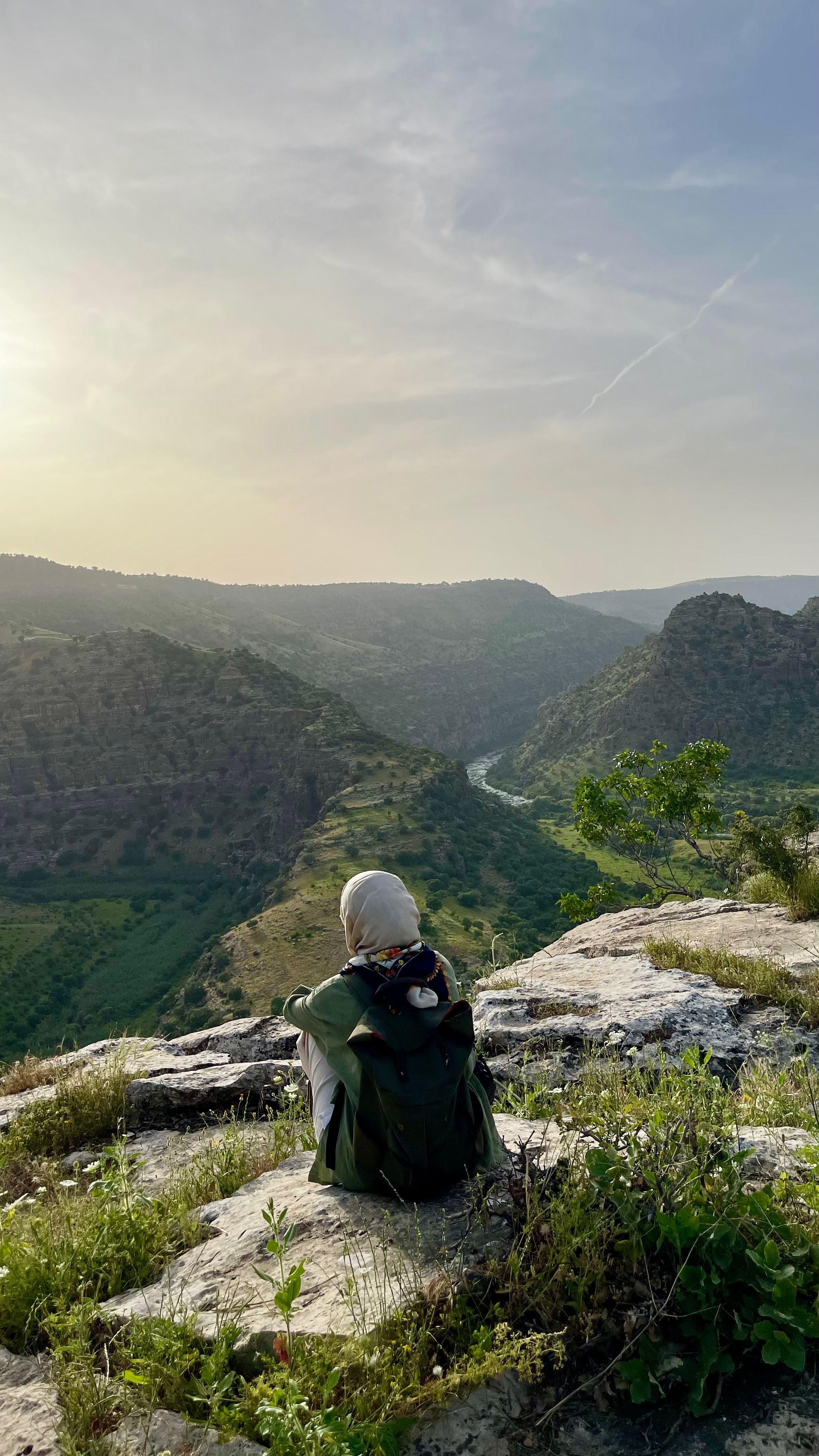 A woman in a hijab sits on a hilltop, enjoying a peaceful sunset over a lush valley.