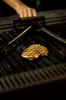 Close-up of a juicy grilled chicken breast on a barbecue grill with tongs. Dark, smoky ambiance.