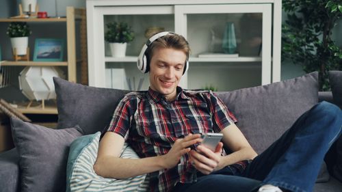 Free Man in plaid shirt relaxing on sofa with headphones and smartphone indoors. Stock Photo