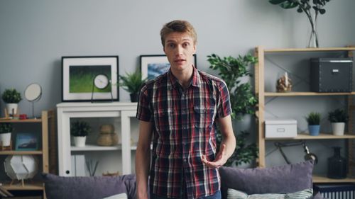 Free Young man standing in a modern living room with decorative shelving, plants, and framed art. Stock Photo