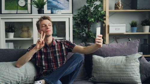Free A young man sitting on a sofa indoors, taking a selfie with a smartphone and making a peace sign. Stock Photo