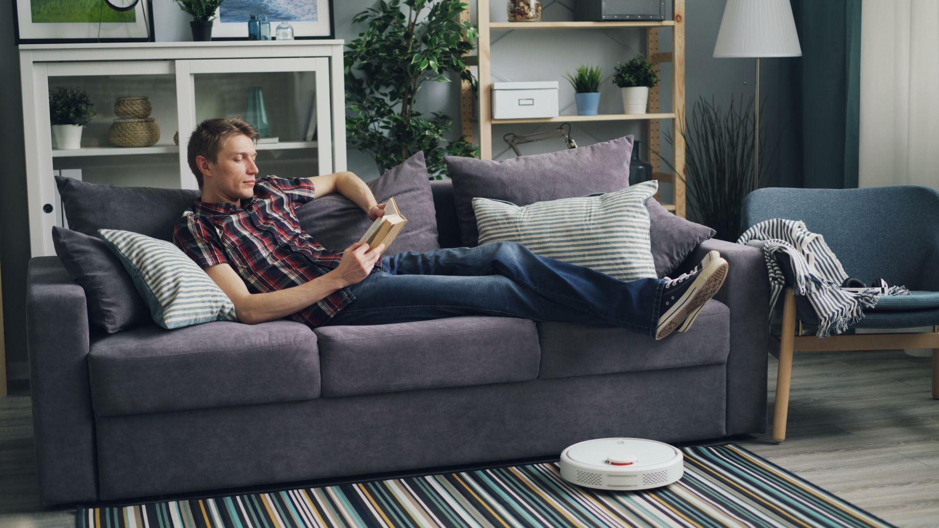 Man Lying on a Sofa and Reading a Book with a Cleaning Robot Vacuuming the Floor