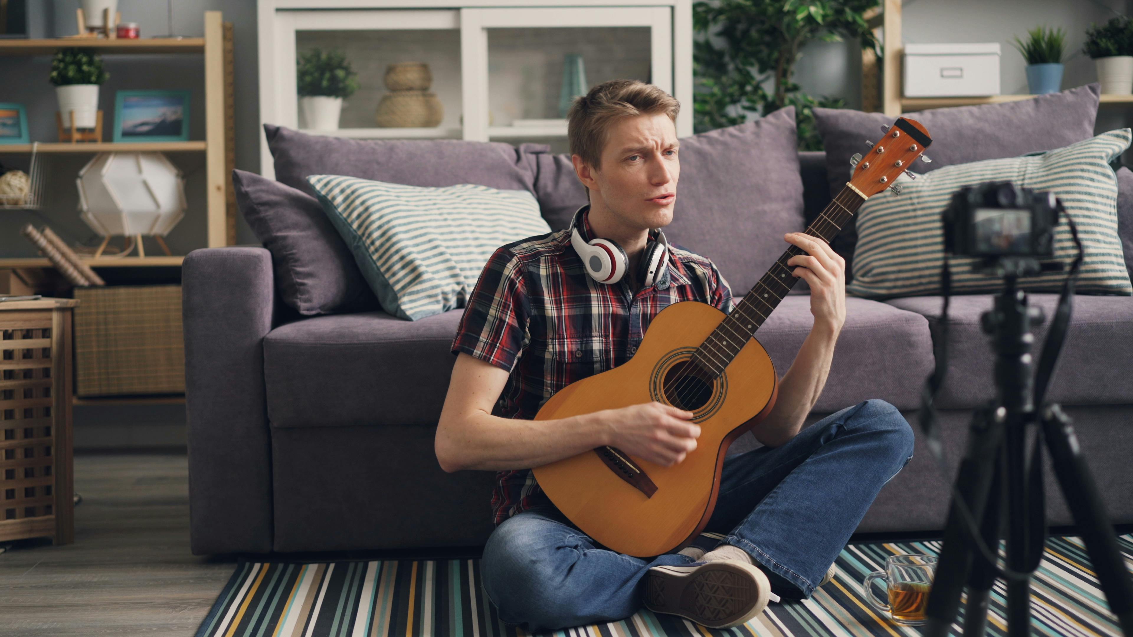 man sitting on a floor cross legged and playing the guitar to the camcorder