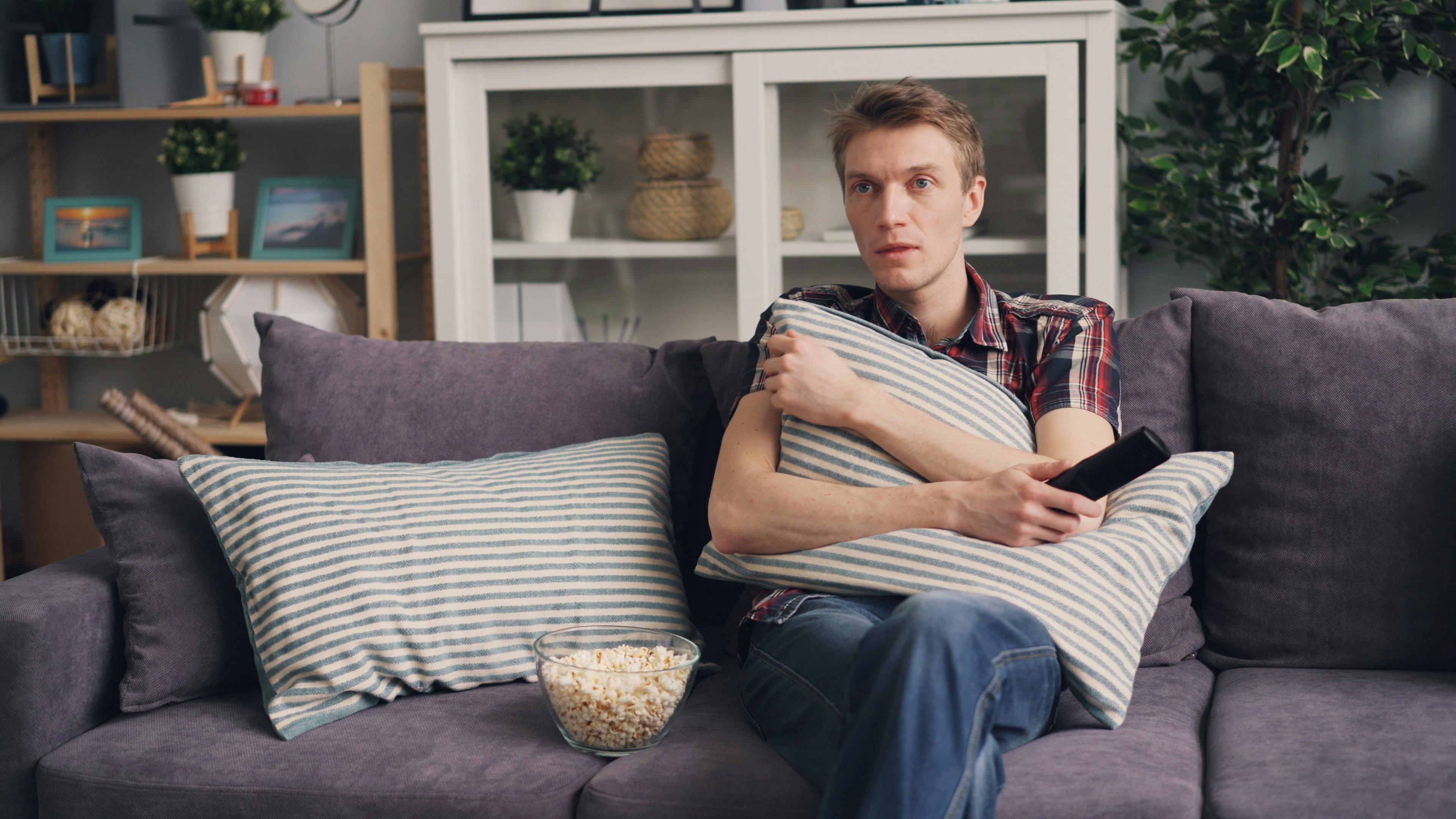 A man sits on a sofa holding a pillow while watching TV, with a bowl of popcorn beside him.