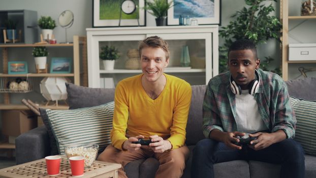 Two friends playing video games together on a sofa, one holding a controller and the other wearing headphones.