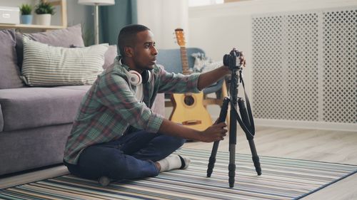 Free Young man setting up a camera on a tripod in a cozy living room setting. Stock Photo