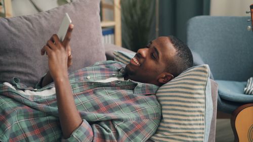 Free Man lying on sofa using smartphone, relaxing in a cozy home environment. Stock Photo