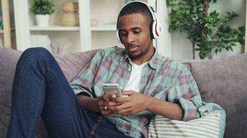 Free Young man wearing headphones sitting on a couch, enjoying music and using a smartphone indoors. Stock Photo