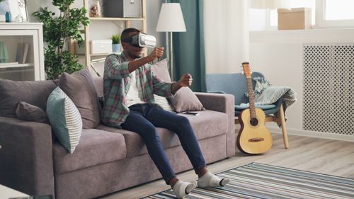 Free Black man using VR headset on sofa at home, surrounded by decor and guitar. Stock Photo