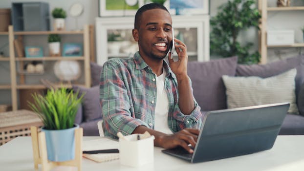 Black man in a casual home office setting, multitasking with a laptop and phone.