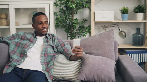 Free A relaxed man smiling while using his smartphone, seated on a cozy couch indoors. Stock Photo
