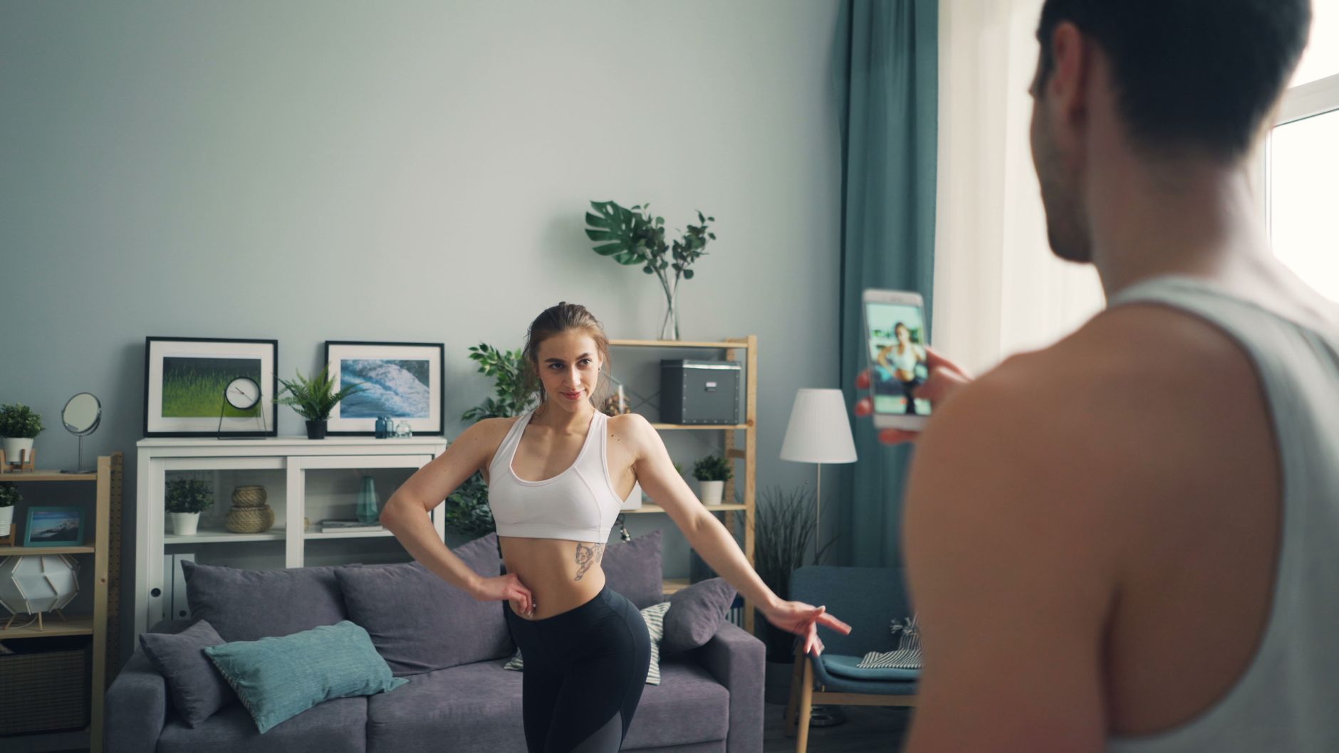 A young woman in sportswear poses confidently as a man records her with a smartphone in a bright living room.