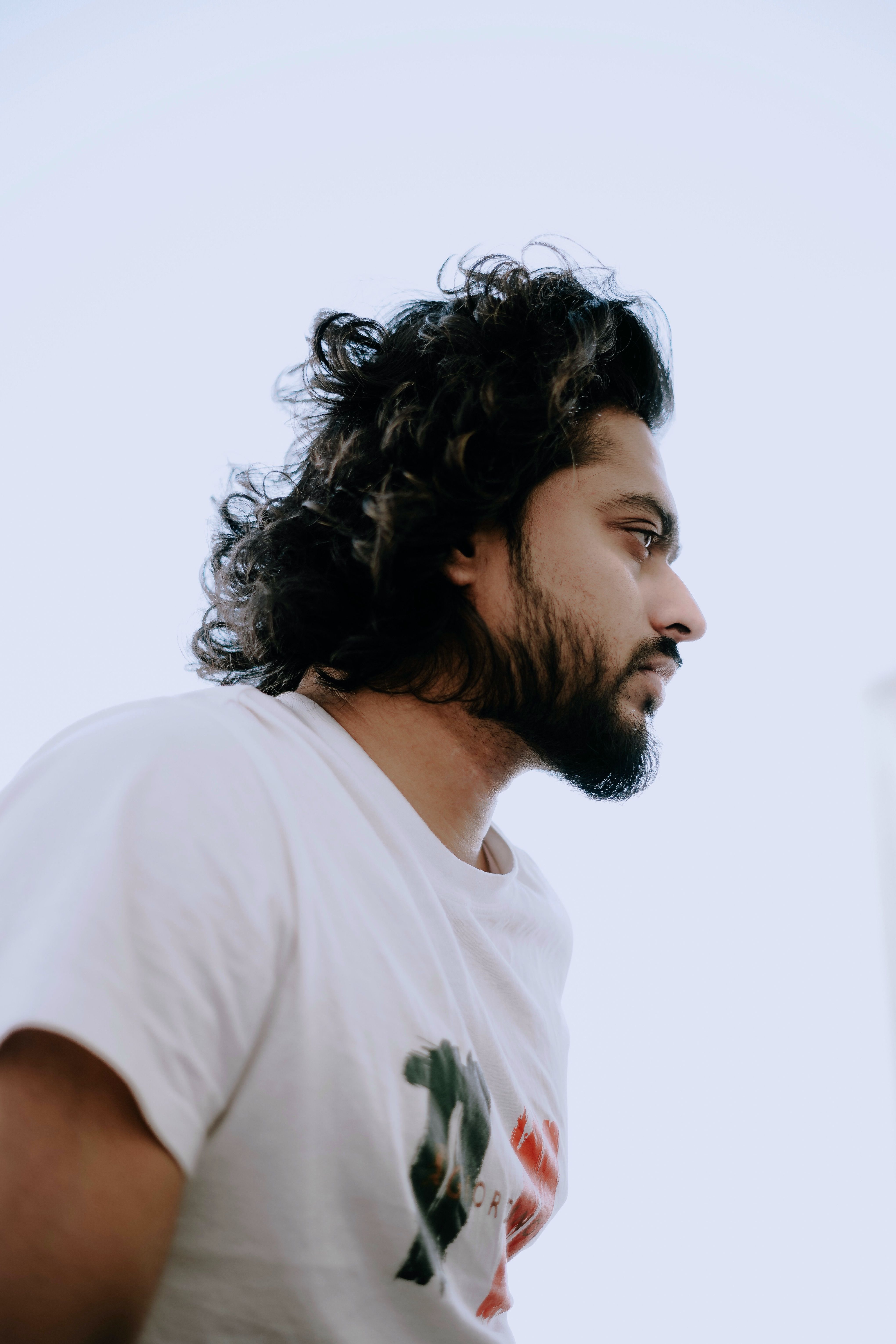Profile view of a bearded man with long hair outdoors, wearing a white t-shirt.
