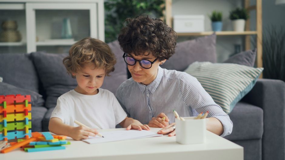 A mother helps her young son with drawing at a table in a cozy living room setting.