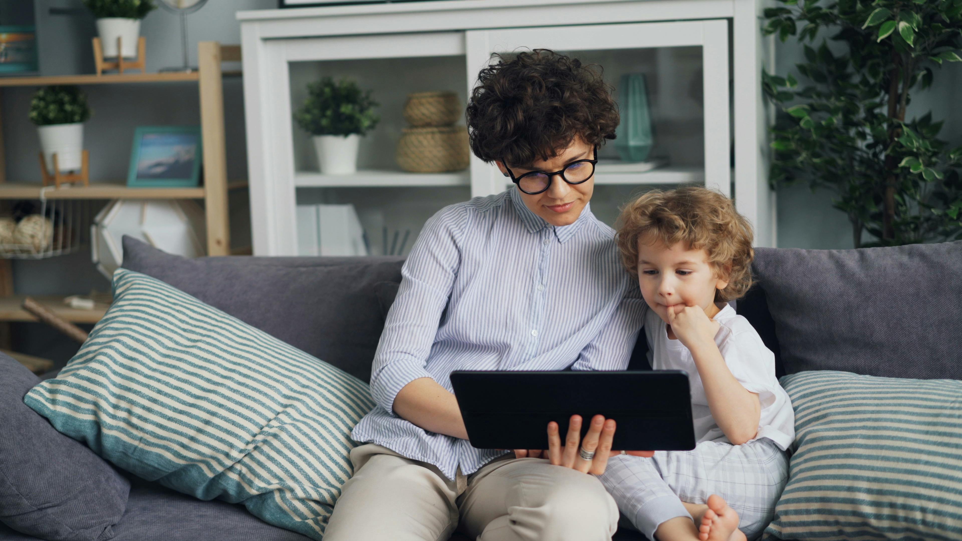 A parent and child sit on a couch engaging with a tablet indoors.