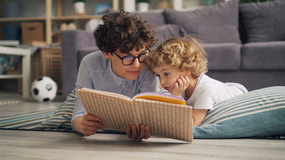 A mother and son lying on the floor reading a book together in a cozy living room setting.