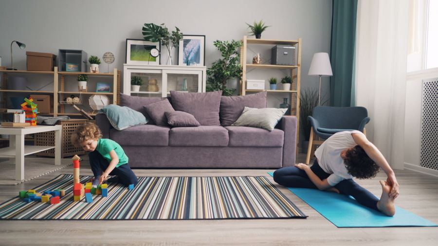 Mother doing gentle stretching on a yoga mat at home while a child plays nearby