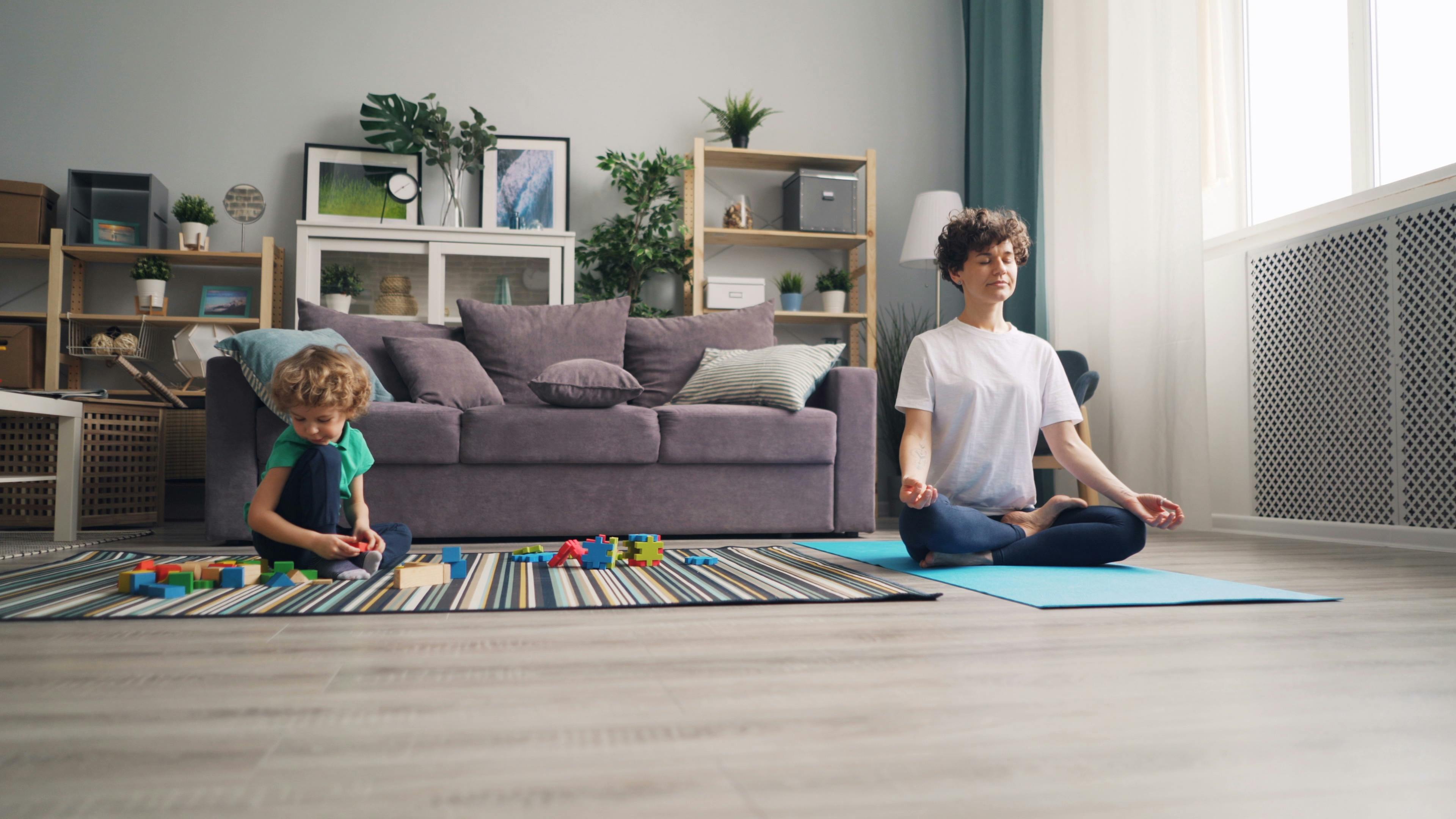 Free A mother meditates on a yoga mat while her son plays with blocks in a cozy living room setting. Stock Photo