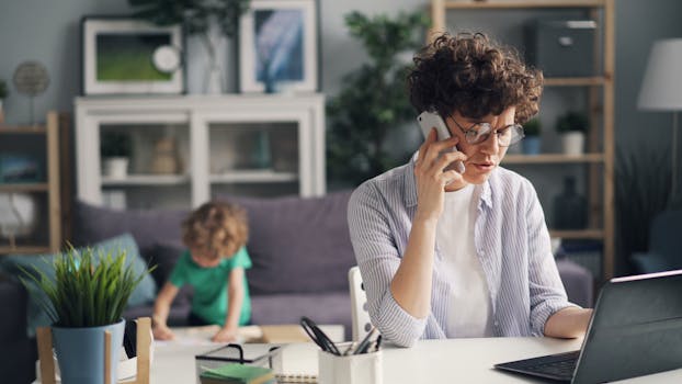 A mother multitasking with a laptop and phone while her child plays in the background at home.