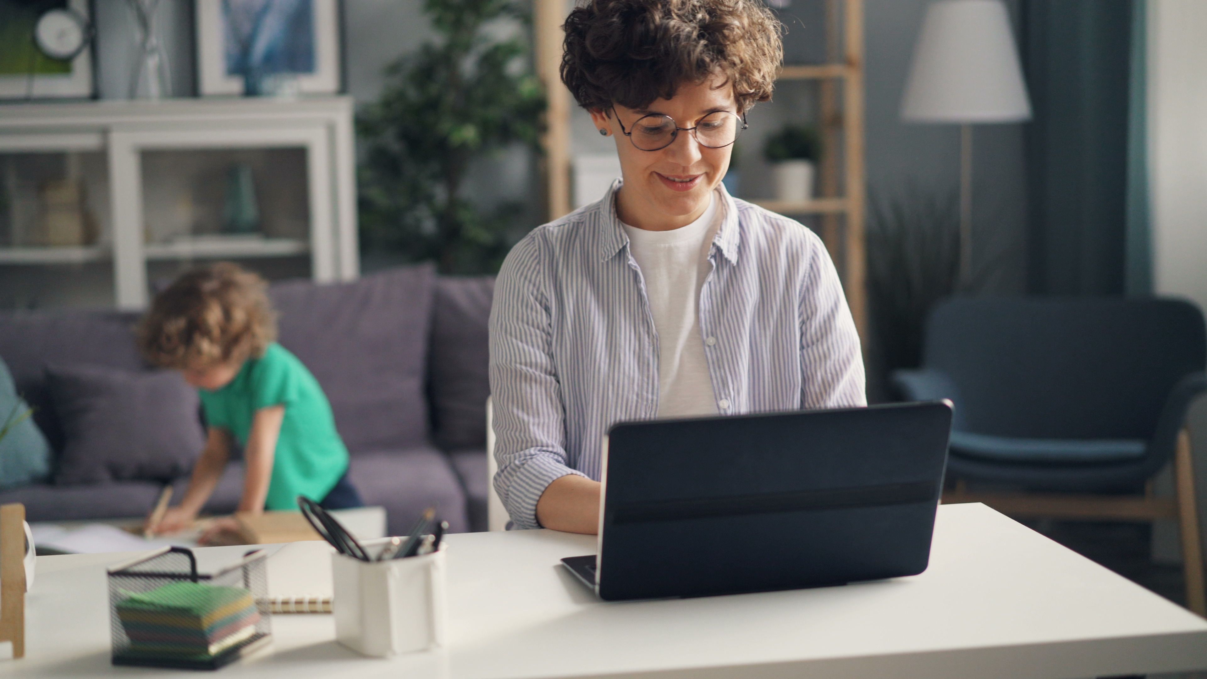 This photo shows a woman working on a laptop while her son studies in the background.