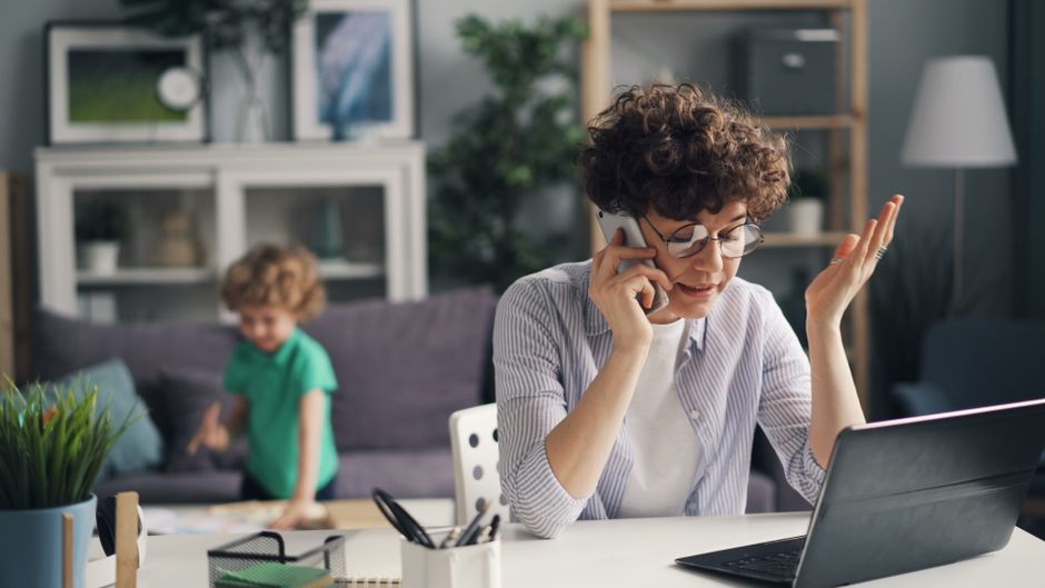 A woman multitasking with a phone and laptop while her child plays in the background.