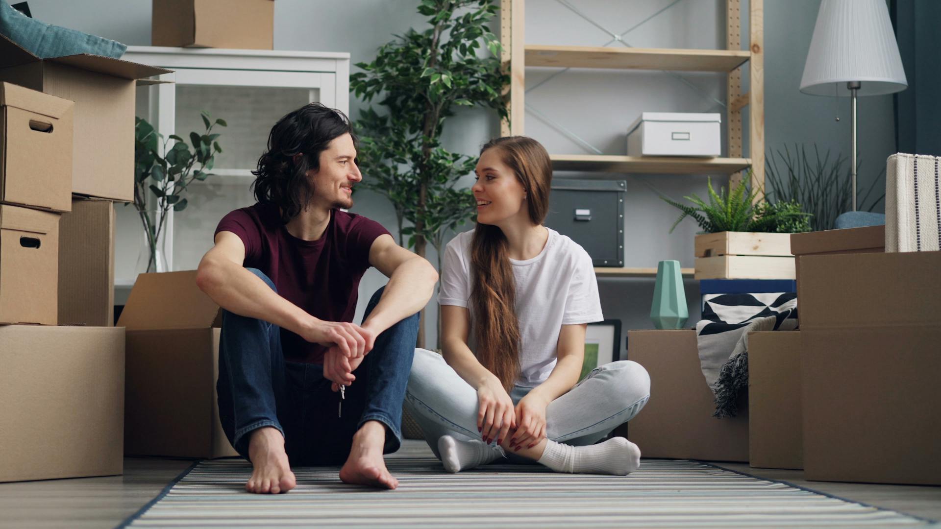 Smiling couple sitting on the floor amidst moving boxes, embracing new beginnings in their new home.