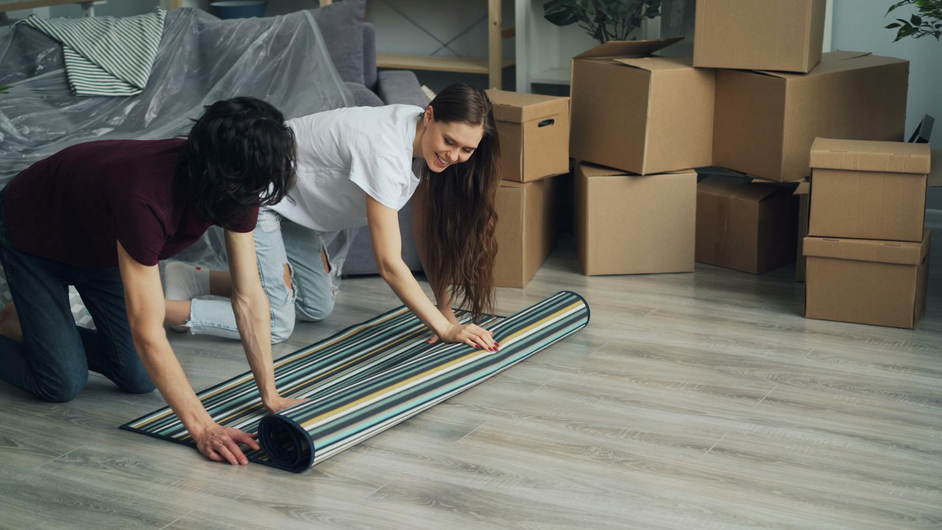 Couple smiling as they unroll carpet surrounded by moving boxes in their new home.