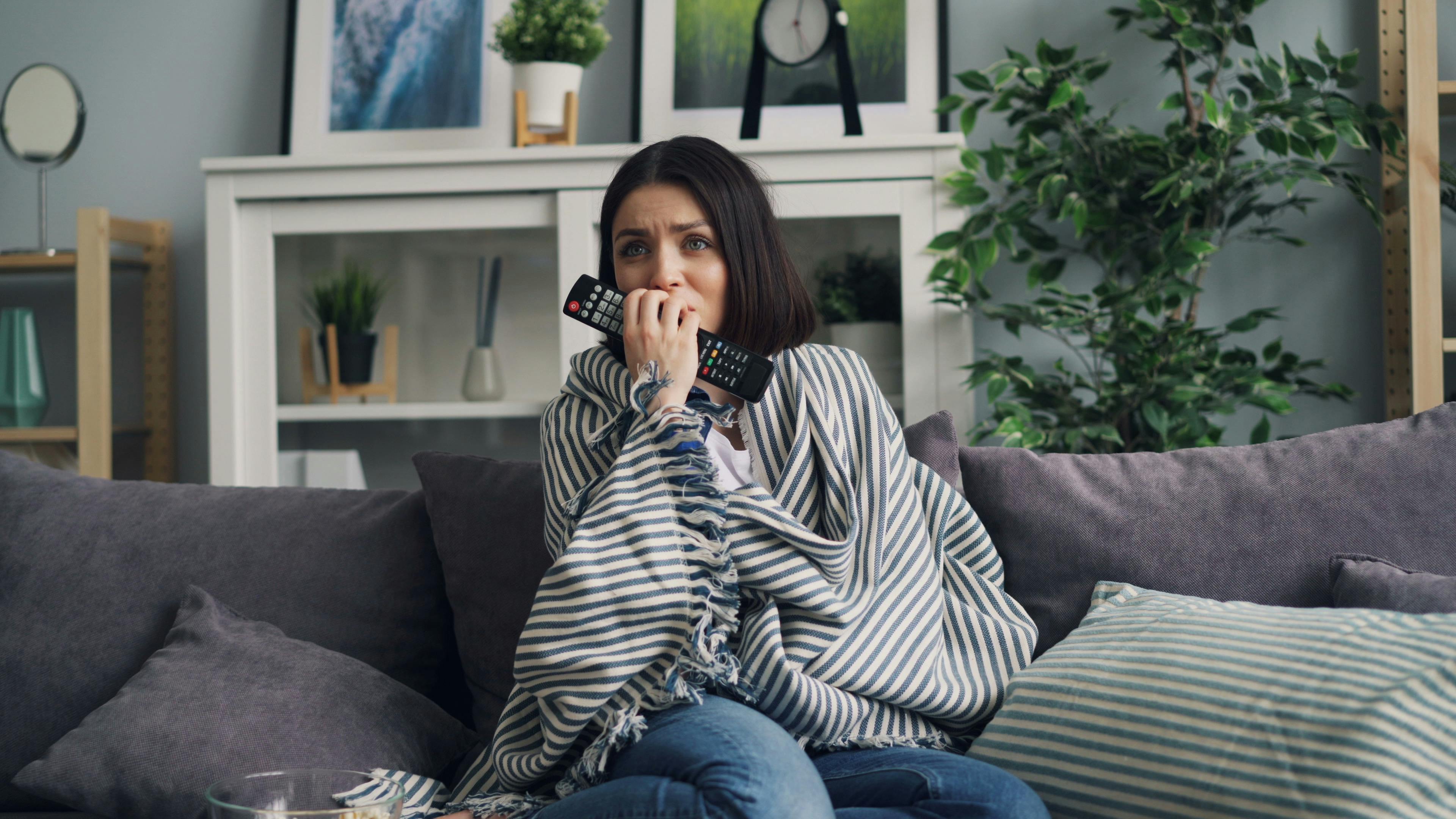 Anxious woman wrapped in a blanket watching TV at home with a remote in hand.