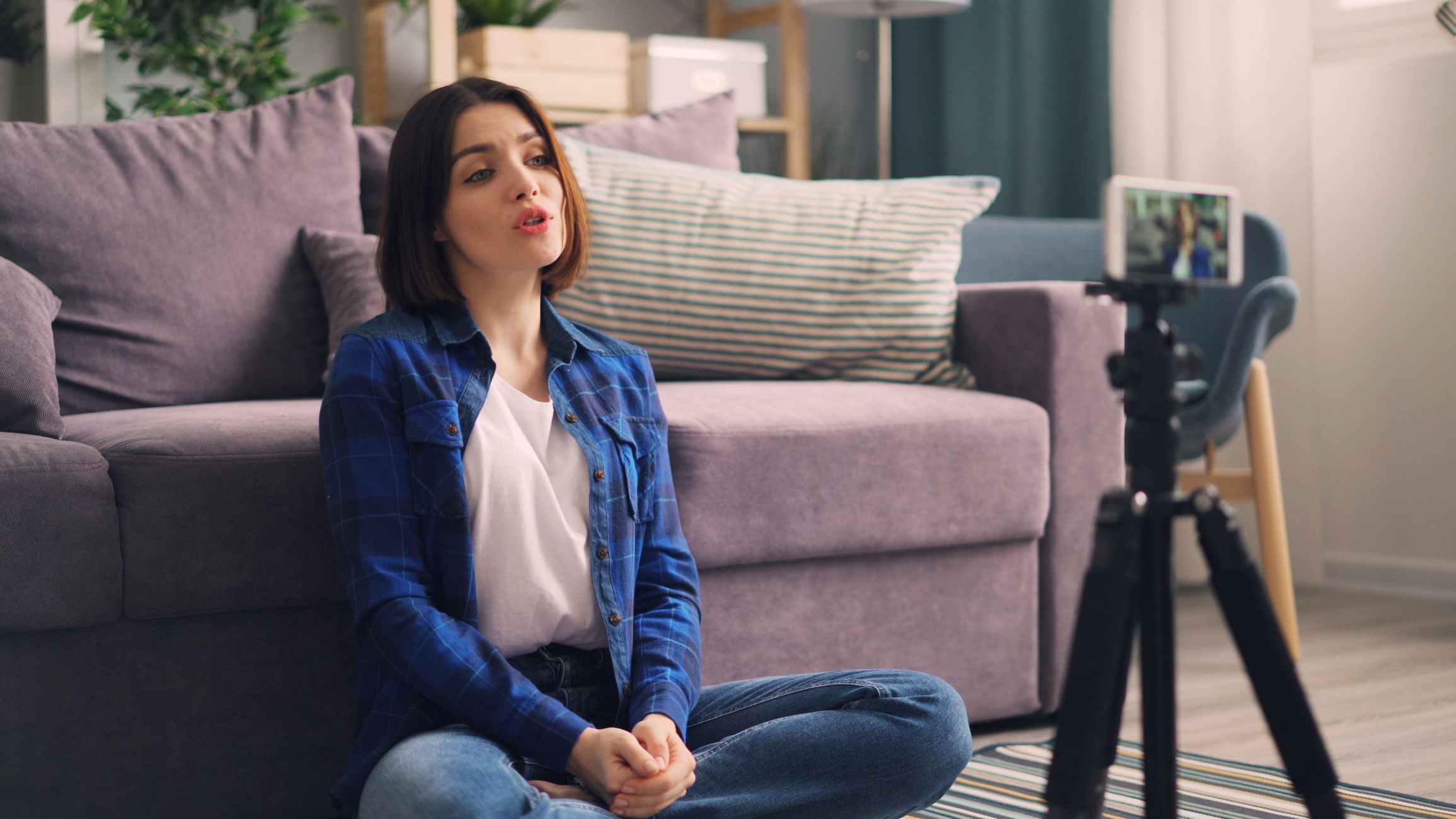 Adult woman recording video indoors on a smartphone, sitting cross-legged on the floor.