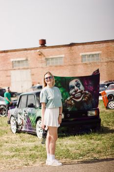 Woman standing at a car show featuring a car with a vibrant Joker-themed hood design.
