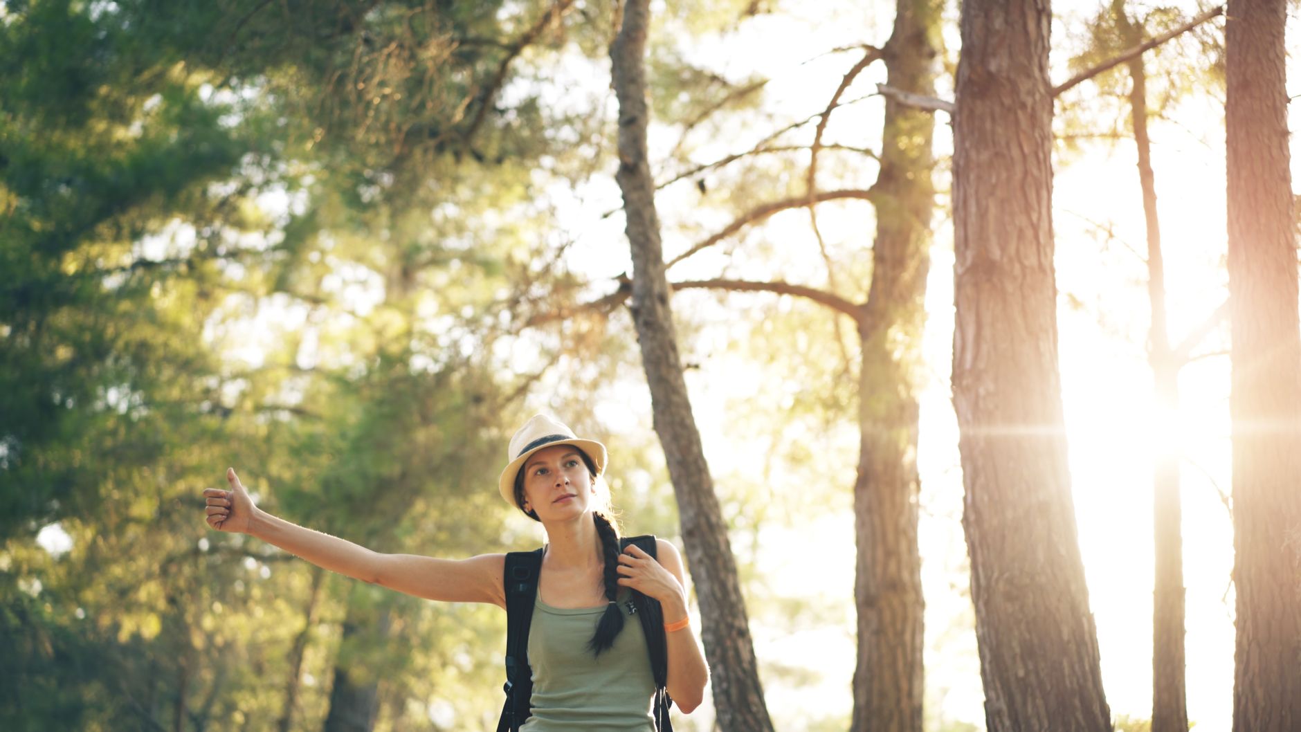 nmn for midlife energy crash women — A woman hitchhikes in a sunlit forest, with a backpack and hat, enjoying the great outdoors.