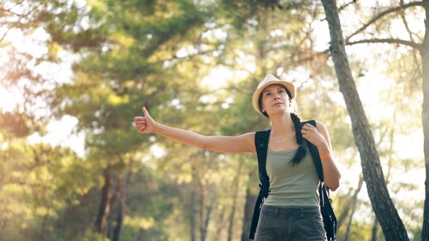 A woman with a backpack hitchhikes on a sunny day in the forest, wearing a hat.