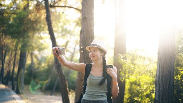 A woman with a hat and braids taking a selfie in a sunlit forest during sunset.