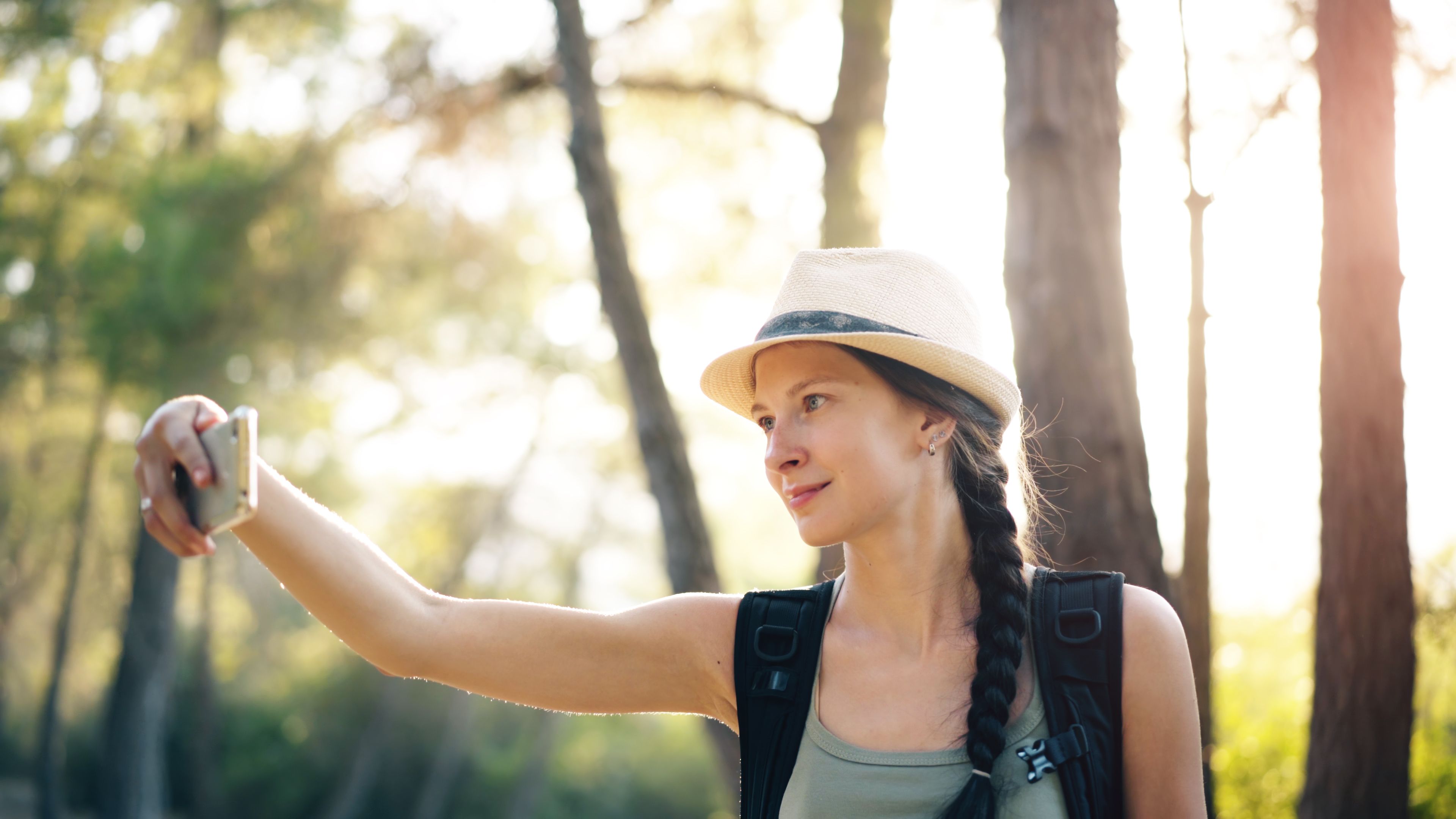 Woman with a braid and hat taking a selfie in a sunlit forest during a hike.