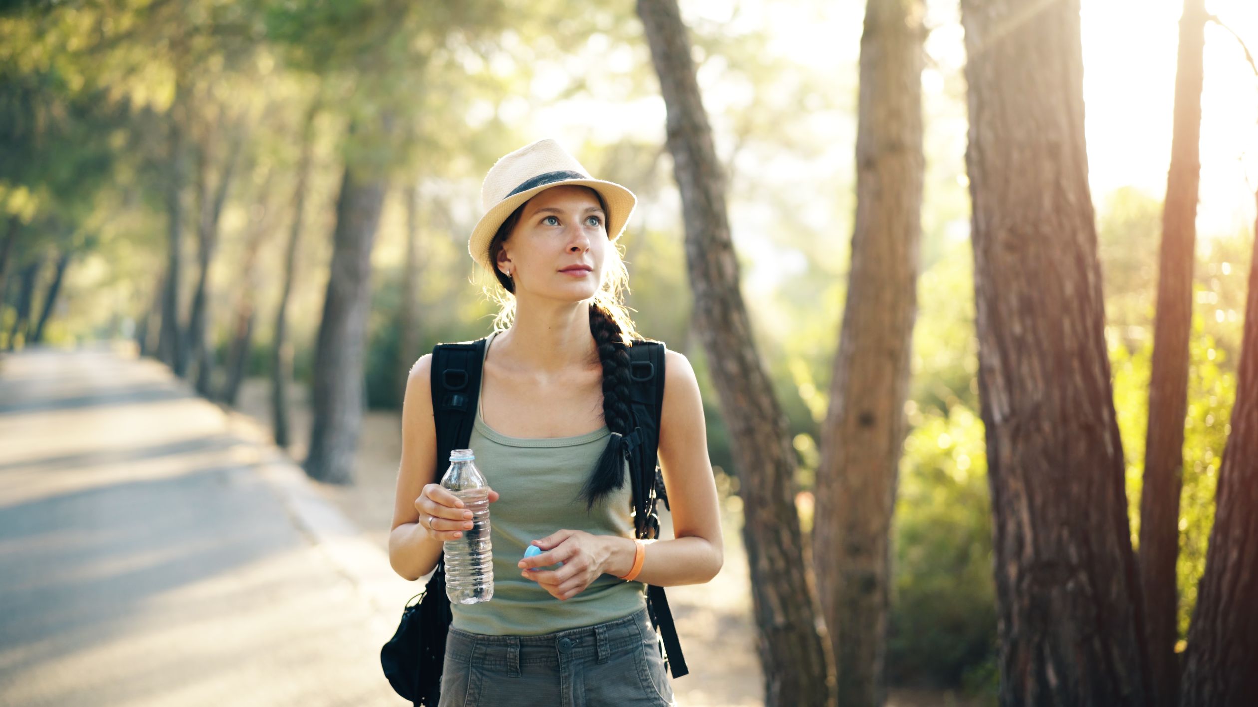 Woman with braid and hat hiking in a forest, holding a water bottle