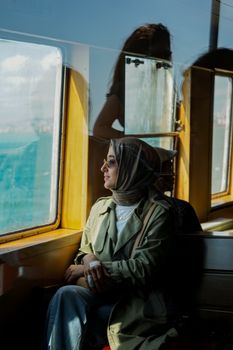 Woman in headscarf sitting by ferry window, gazing at sea view.