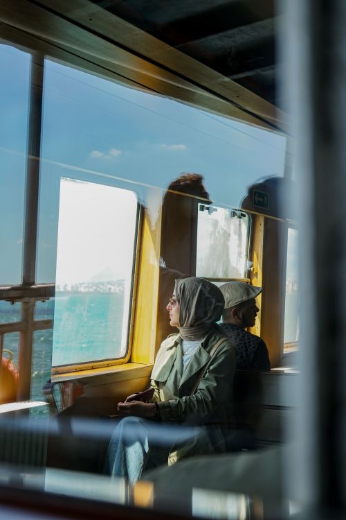 Free A thoughtful woman in a headscarf looks out from a ferry window, capturing a serene travel moment. Stock Photo