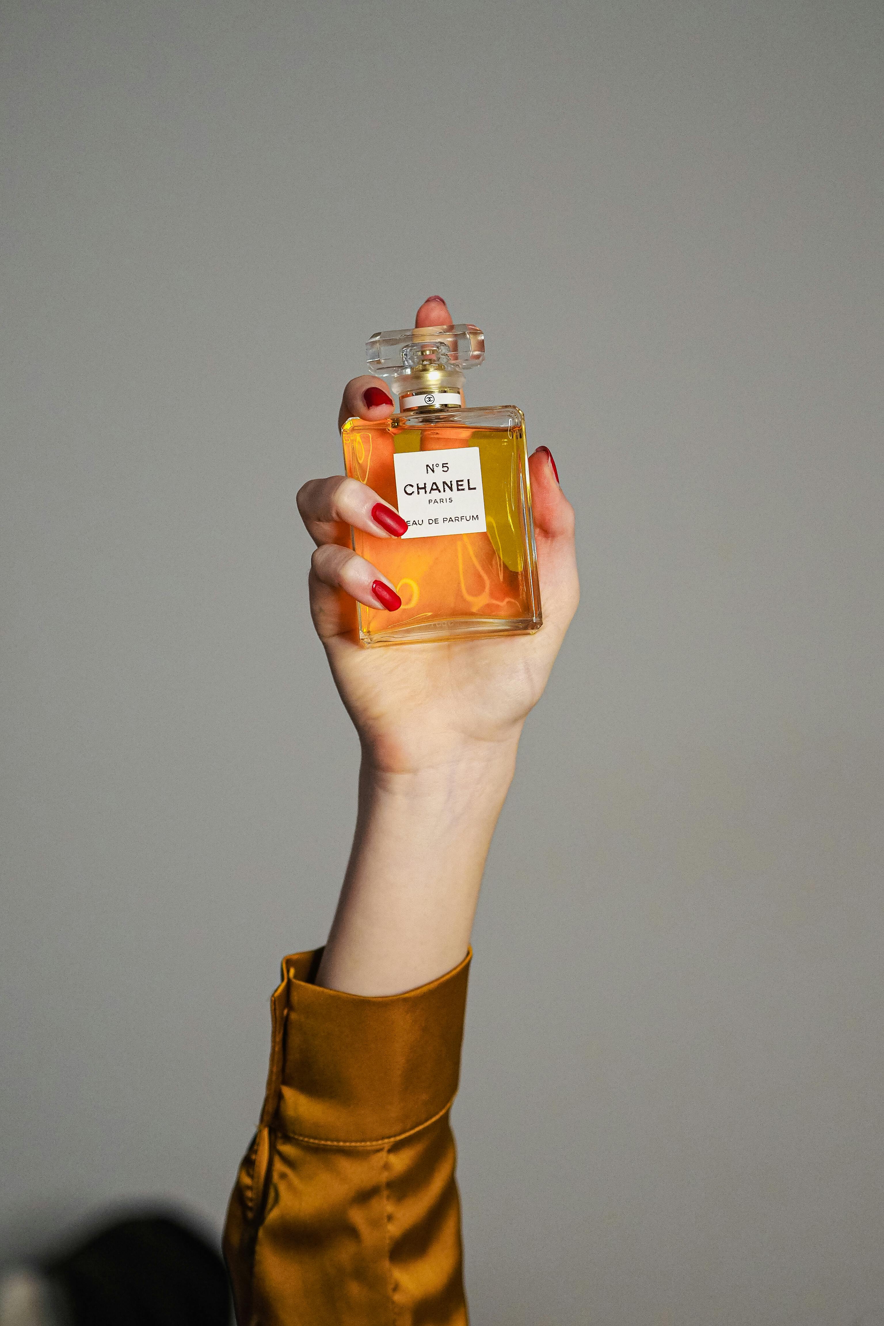 A hand with red nails holds a Chanel No. 5 perfume bottle against a simple gray background.