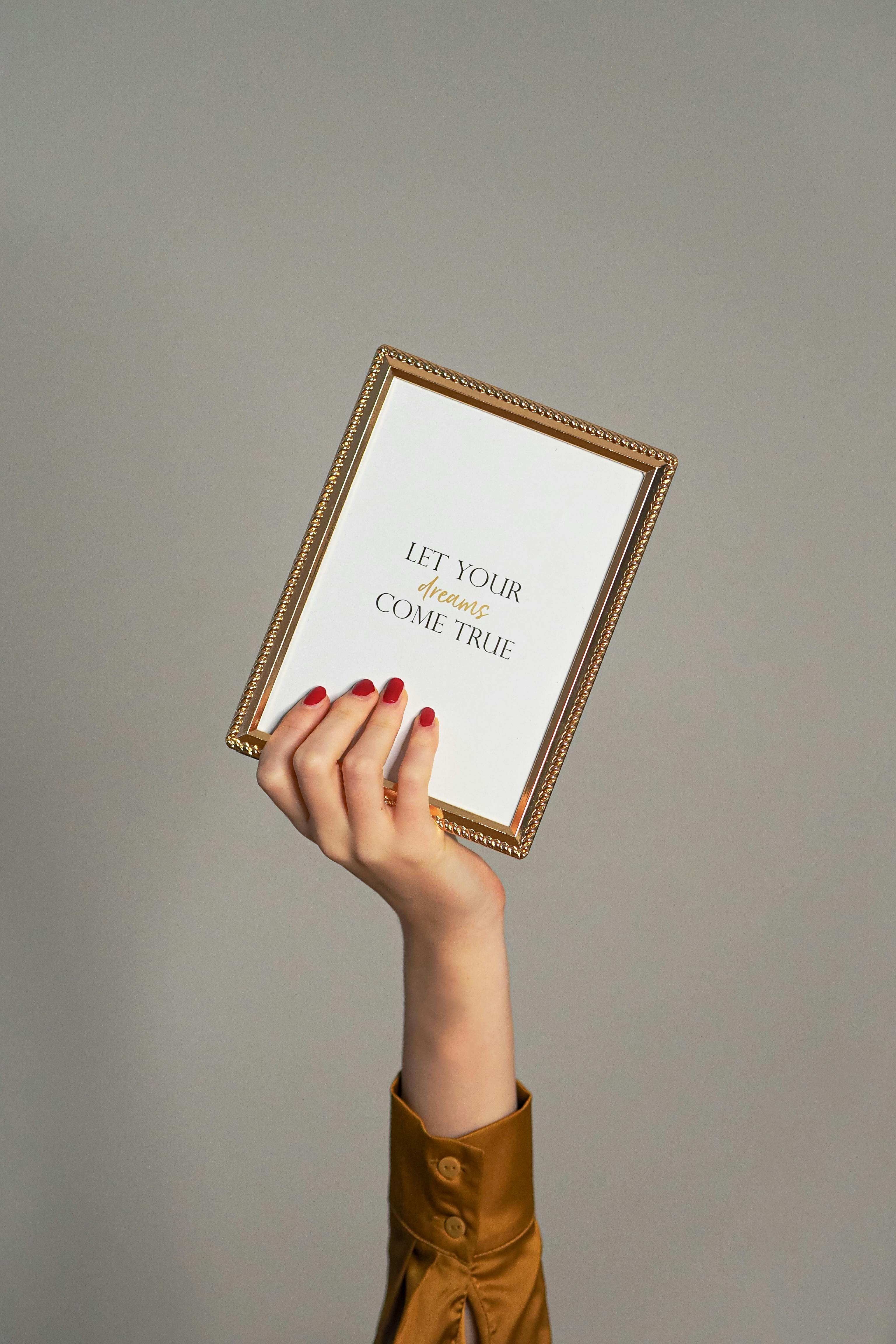 A framed quote saying 'Let Your Dreams Come True' held by a hand.