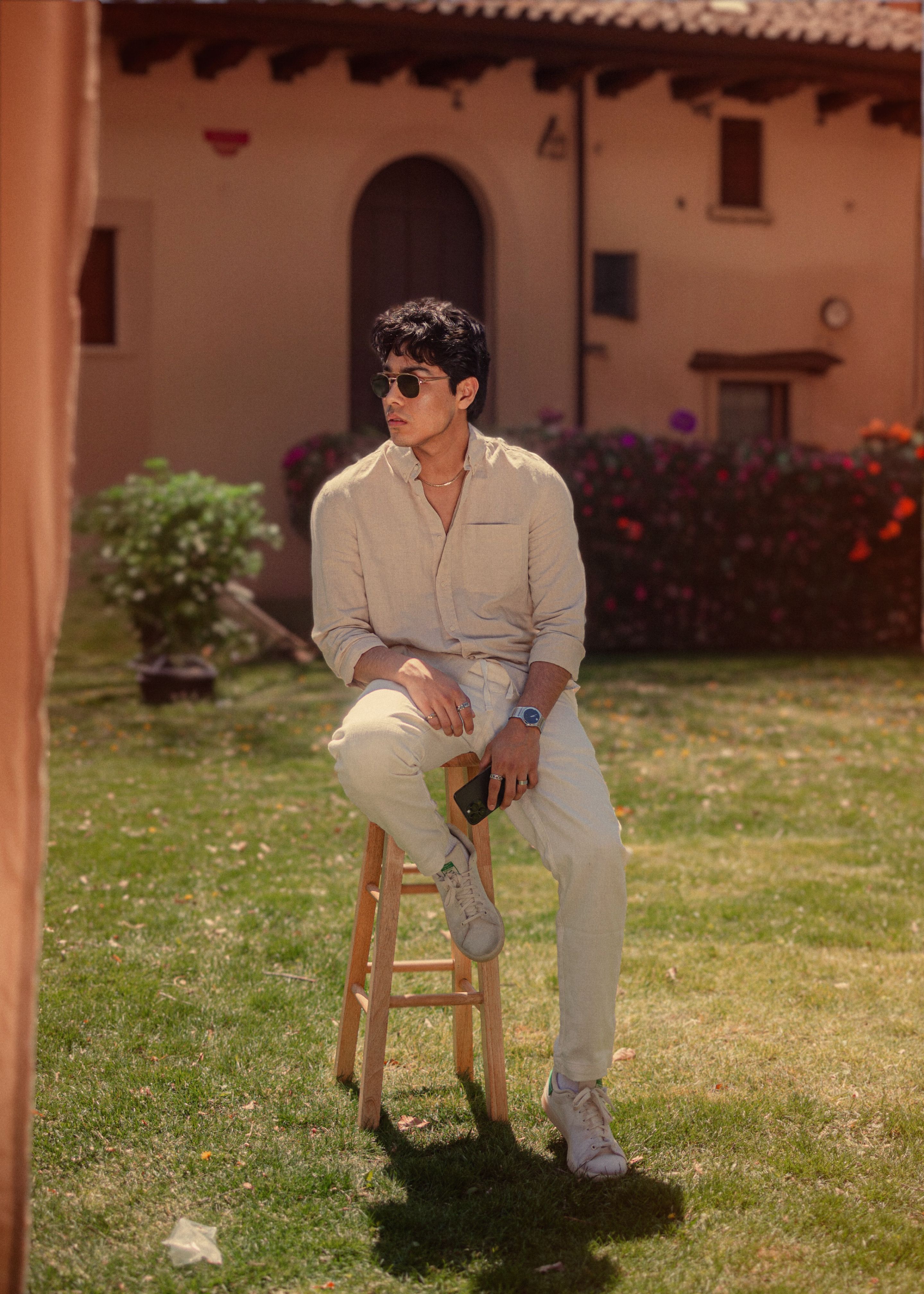 A fashionable man in sunglasses sits on a stool in a sunlit garden, wearing casual beige attire.