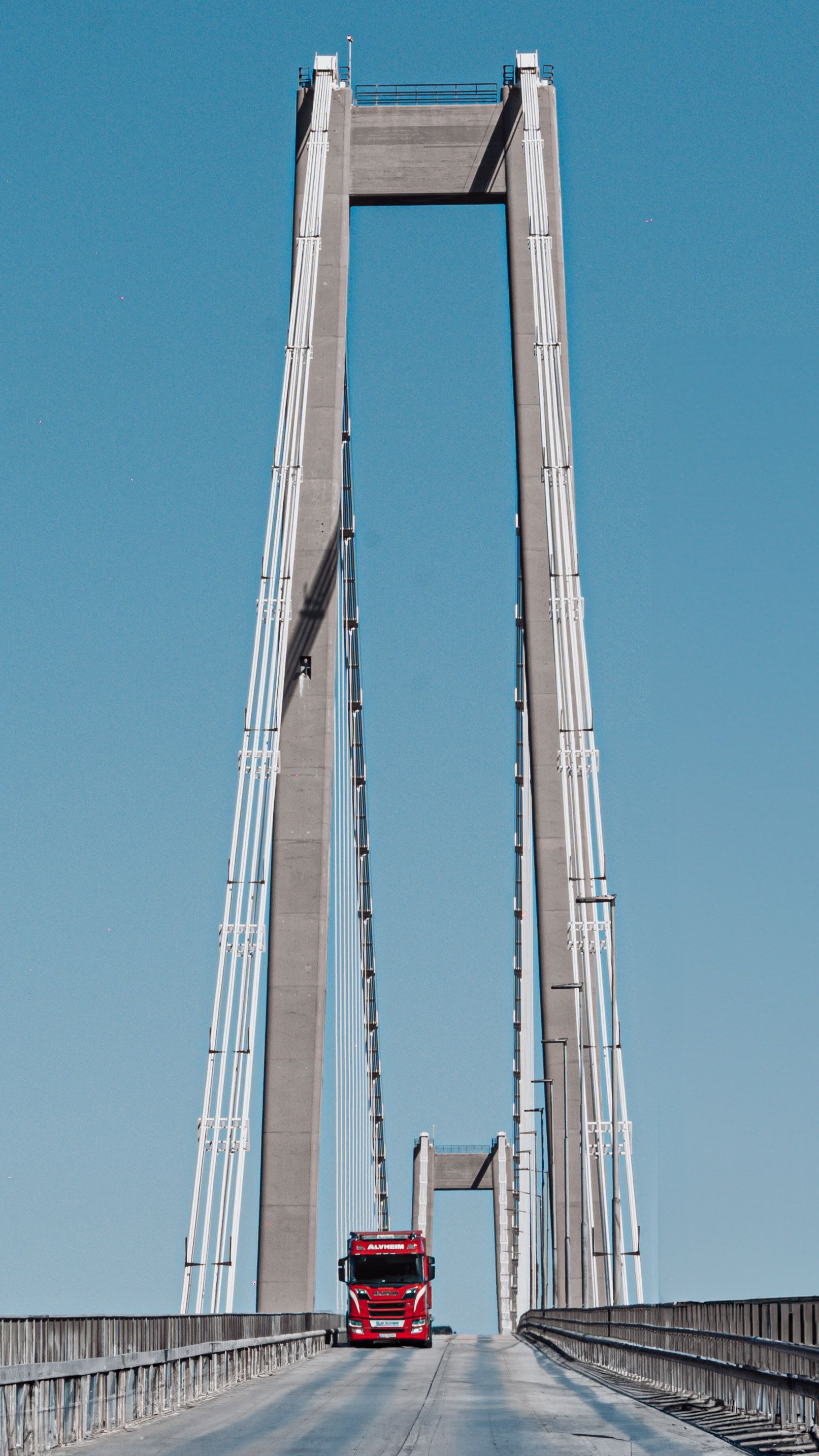 A red truck drives over a tall suspension bridge against a clear blue sky.