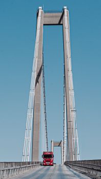 A red truck drives over a tall suspension bridge against a clear blue sky.